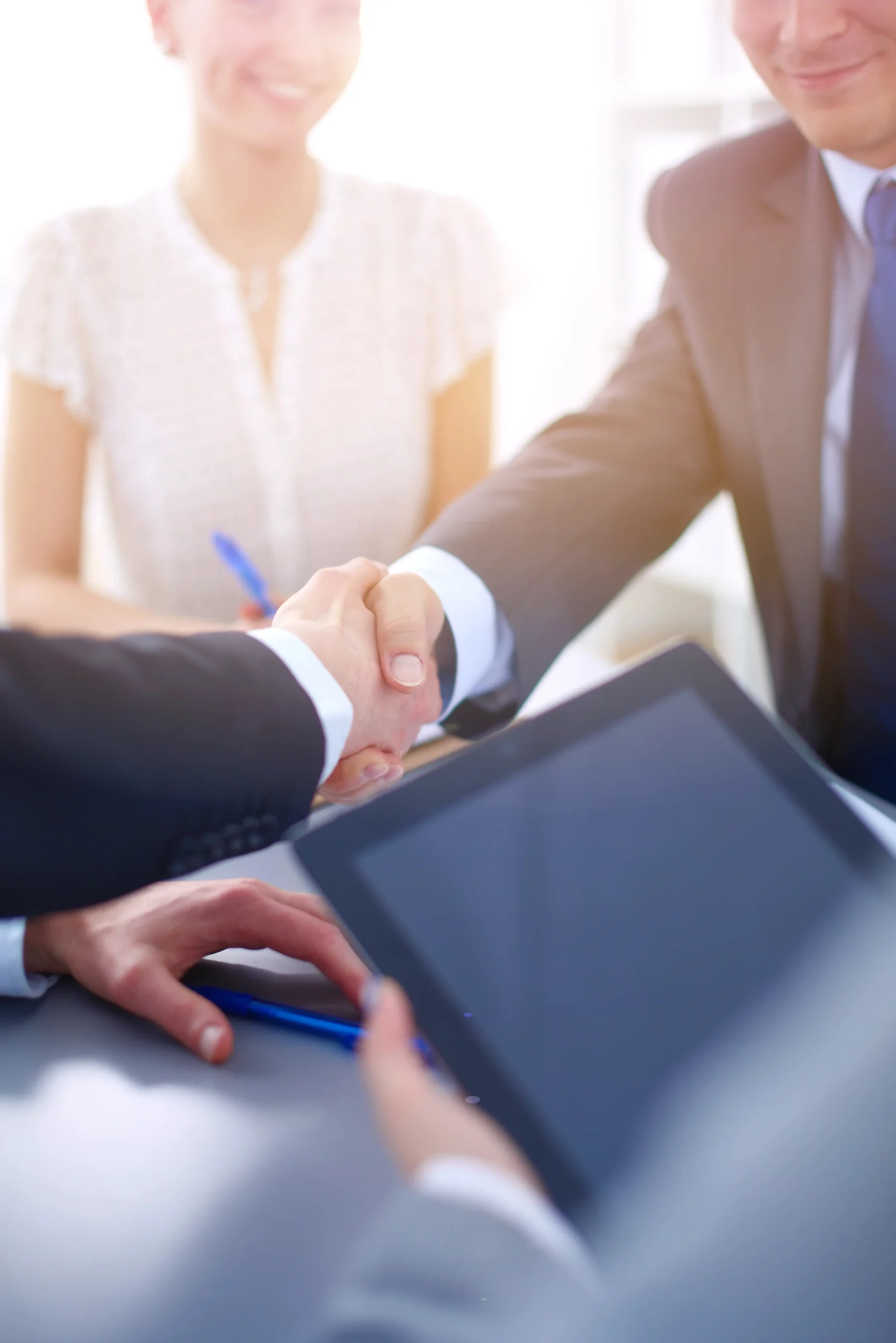 Business professionals in formal attire shaking hands over a desk, conveying agreement, with a tablet in the foreground and a smiling woman nearby